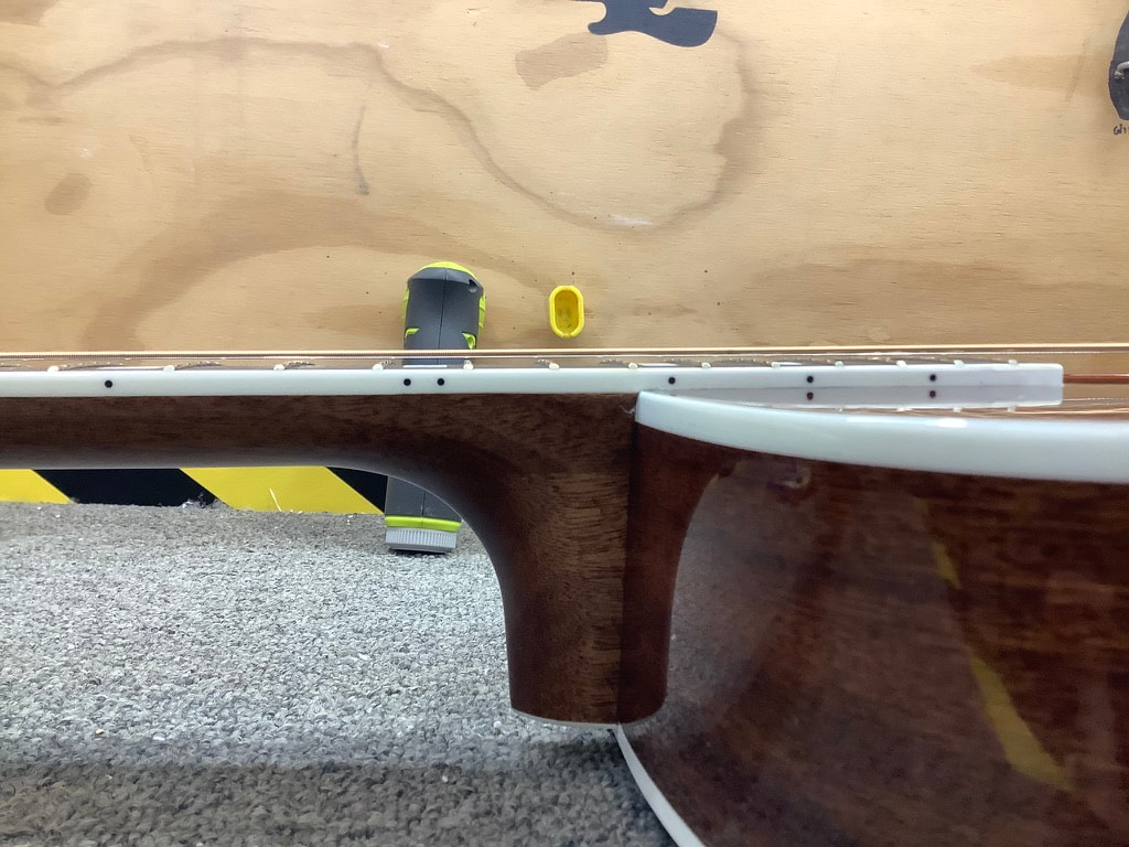 Vista lateral del mástil y el cuerpo de una guitarra acústica desde el traste nueve al veinte. Se aprecian las cuerdas bajas, el tacón y el hombro de la guitarra en primer plano. 