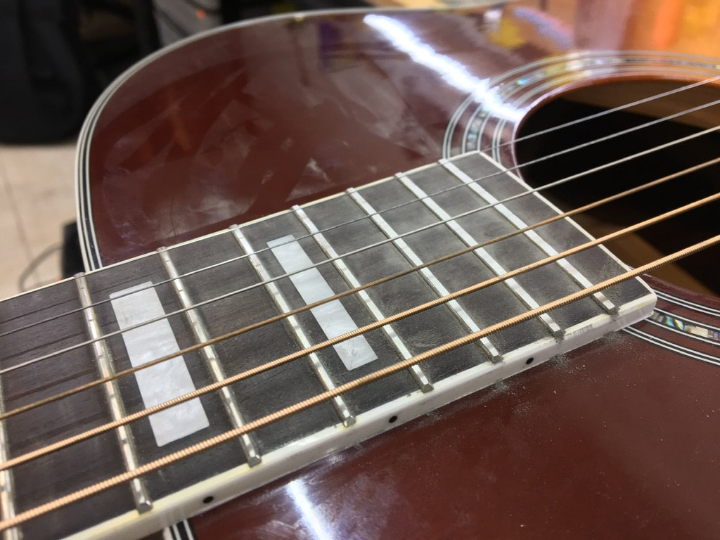 Foto de diapasón de guitarra acústica color oscuro con ingrustaciones rectangulares, con los trastes lijados y las cuerdas instaladas en una guitarra acústica con terminado tobacco burst